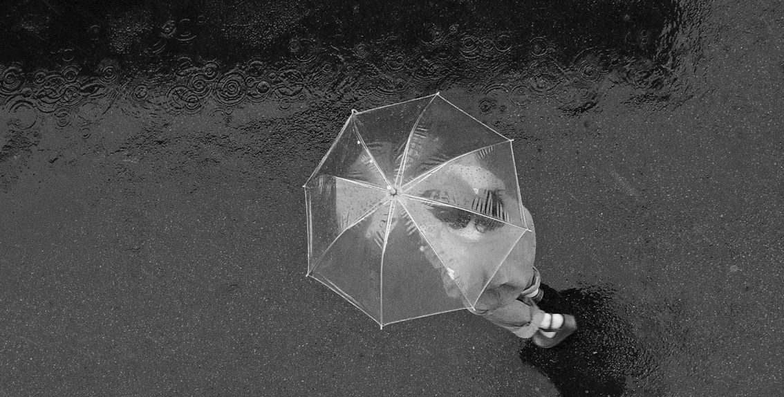 Uma pessoa vista de cima, caminha na chuva com um guarda-chuva transparente. No asfalto, as gotas d´água desenham círculos nas poças que se formaram.